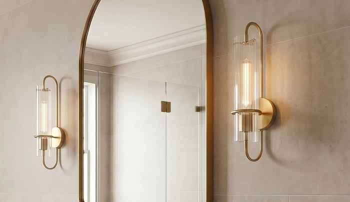 Two modern Cami wall sconce light fixtures with a brushed gold metal frame and clear glass shades, mounted on a beige tiled wall next to a large arched mirror showing its reflection.