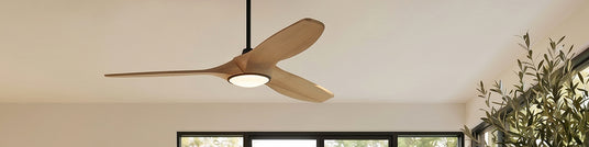 Natural wood, modern-designed three-blade smart ceiling fan with light in a family room with off-white walls and black frame industrial windows.