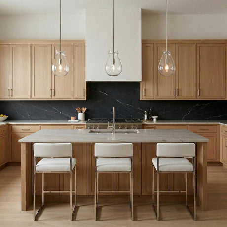 A transitional kitchen features three Emma Pendant Light 7", Polished Nickel glass fixtures over a marble island with light wood cabinetry and a black marble backsplash.
