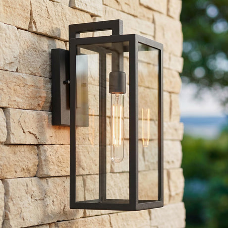 A rectangular modern home exterior with a textured black Sam Outdoor Wall Light, Medium 18" glowing on a tan stacked stone wall beside blurred green landscaping at twilight.