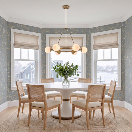 A coastal dining room featuring the Castell 6 Globe Round Chandelier 36 in Aged Brass over a white pedestal table with woven rattan chairs and blue wallpaper.