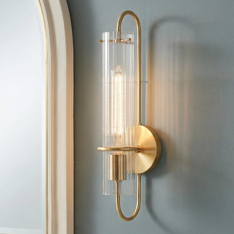 A modern bathroom featuring the Camie Wall Sconce 20", Aged Brass with ribbed glass mounted on a blue-gray wall beside a large white arched mirror.