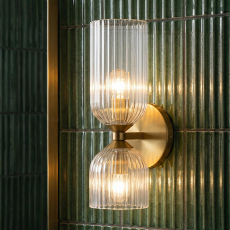 A modern bathroom featuring the Elle Wall Sconce, Brushed Brass and Clear Glass, mounted on vertical green fluted tile beside a gold mirror frame.
