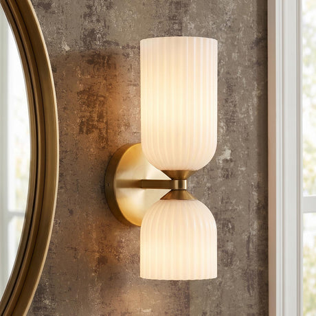 A contemporary bathroom with an Elle Wall Sconce, Brushed Brass and Milk Glass featuring ribbed shades, mounted on textured wallpaper beside a round gold mirror and window.