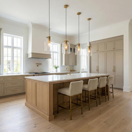 A transitional kitchen featuring four glass pendants with brushed brass accents over a large white marble island accompanied by taupe cabinetry and four upholstered bar stools.