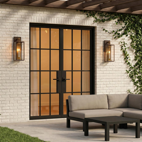 A modern farmhouse patio featuring the Orin Outdoor Wall Light in bronze with vintage brass accents flanking black glass doors on white brick at twilight with ivy.