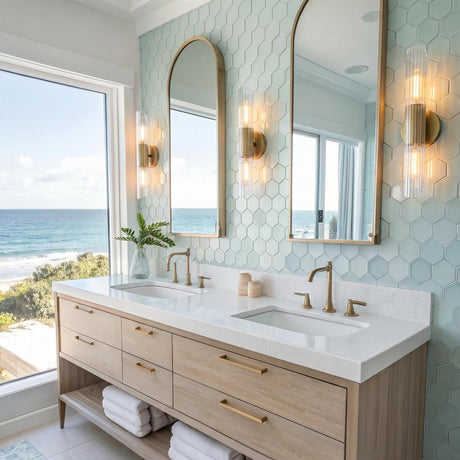 A coastal bathroom with three aged brass and ribbed glass wall sconces flanking arched mirrors over a light wood vanity and mint green hexagon tiles.