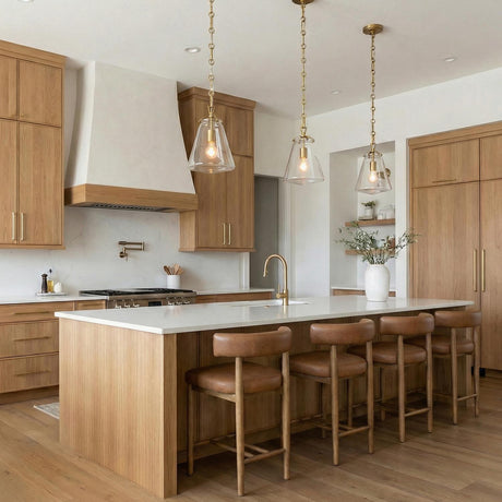 A transitional kitchen features three Addison Small Pendant 8", Brass fixtures with glass shades over a white marble island and light wood cabinetry with leather barstools.