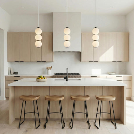 A minimalist kitchen featuring three Shai LED Pendant 7 inch Matte Black and Opal Glass pendants over a white island with light oak cabinetry and four wooden stools.