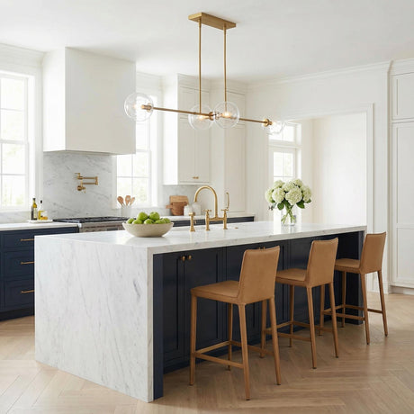 A transitional kitchen features a Gurt 4 Light Linear Pendant 48", Aged Brass with clear glass globes over a marble waterfall island and navy cabinets.
