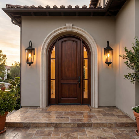 A Mediterranean stucco entry featuring two matte black Asbury Outdoor Wall Lights flanking an arched mahogany door at dusk, with a stone tile porch and greenery.