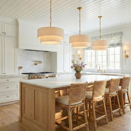 A coastal kitchen featuring three beige linen drum pendants with brushed brass accents over a white marble island and natural wood cabinetry under a shiplap ceiling.