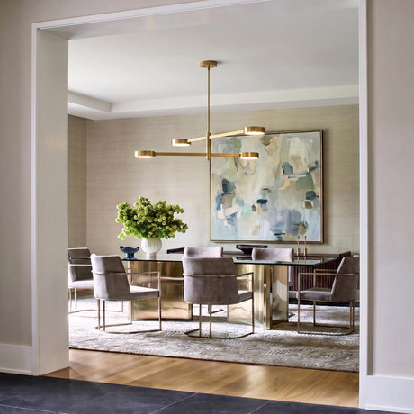 A contemporary dining room with a brushed brass linear chandelier suspended over a glass-top table with abstract wall art, grey upholstered chairs, and grasscloth wallpaper.