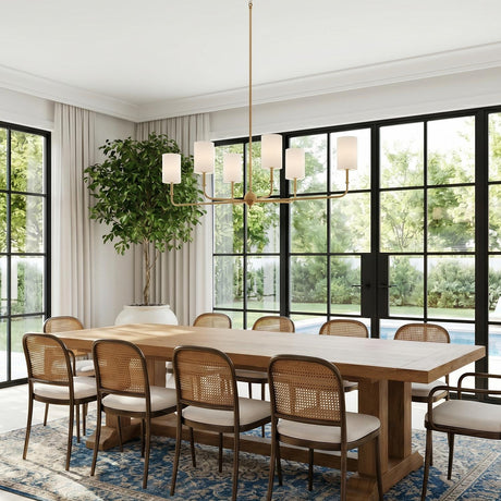 A transitional dining room featuring the Remington 6 Light Linear Chandelier 44", Satin Brass over a long oak table and rattan chairs in front of black-framed windows.