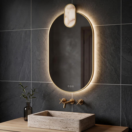 A modern bathroom featuring the Palmer Medium LED Oval Mirror in satin brass and alabaster mounted above a stone vessel sink against dark grey tiles.