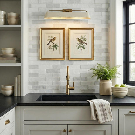 A transitional kitchen features a patina brass and soft sand Henley Picture Light 25" mounted over the sink against a white subway tile backsplash with framed bird prints.