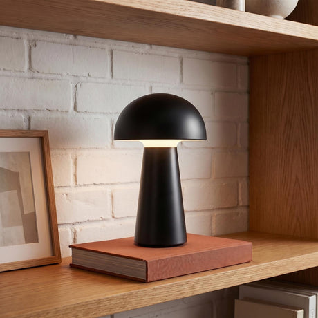 A modern living room featuring the matte black Marnie Rechargeable Table Lamp 9" resting on an orange book atop a light oak shelf with white brick.