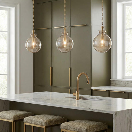 A contemporary kitchen featuring three Arianne aged brass and smoke taupe glass globe pendants suspended over a white marble waterfall island and olive green cabinets.