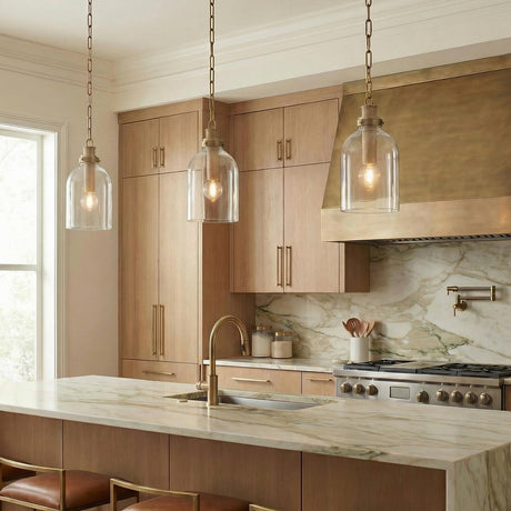 A transitional kitchen features three Midori Small Pendant 9", Patina Brass with Grasscloth Accents over a marble waterfall island beside light wood cabinets and a brass range hood.