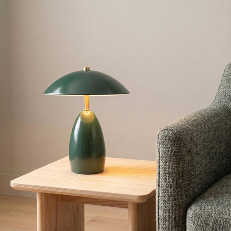 A mid-century living room with the Aubrey Rechargeable Table Lamp 11", Hunter Green in hunter green and brass on a natural wood table beside a gray armchair.