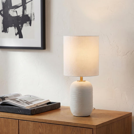 A contemporary living room featuring the Nolette Small Table Lamp 14", Matte White with a ribbed ceramic base and brass neck on a wood sideboard.