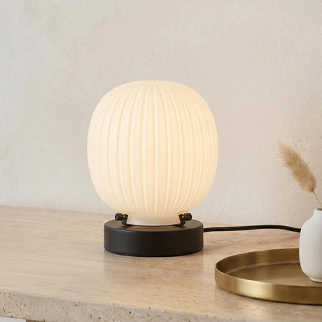 A modern living room featuring the Basile Small Table Lamp with matte black and ribbed milk glass on a travertine console table beside a round brass tray.