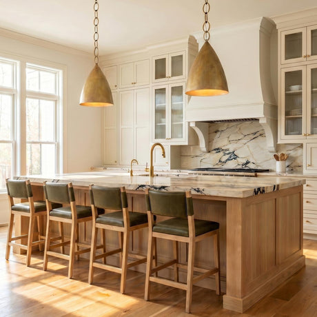 A transitional kitchen with two feathered brass cone pendants hanging over a marble topped wood island featuring cream shaker cabinets and a matching marble backsplash.