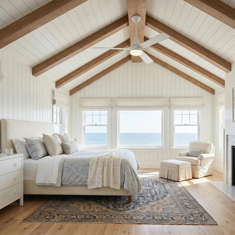 A coastal bedroom with a matte white Serin LED Ceiling Fan 60" along a vaulted beamed ceiling, featuring large windows overlooking the ocean and shiplap walls.