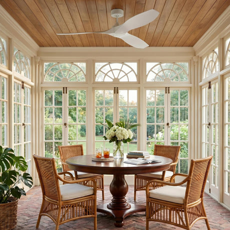 A traditional sunroom with the Heron Outdoor Smart Ceiling Fan 60", Matte White on a wood-paneled ceiling above a brick floor and white window frames.