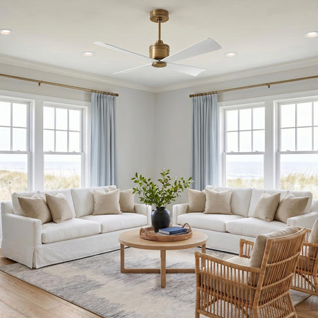 A coastal living room with the Sora Ceiling Fan 44 inch in antique rubbed brass and matte white along the ceiling center and windows overlooking sand dunes.