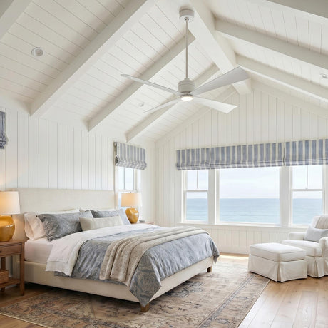 A coastal bedroom with the Sora Ceiling Fan 56", Matte White hanging from a vaulted beamed ceiling above blue patterned bedding and windows overlooking the ocean.