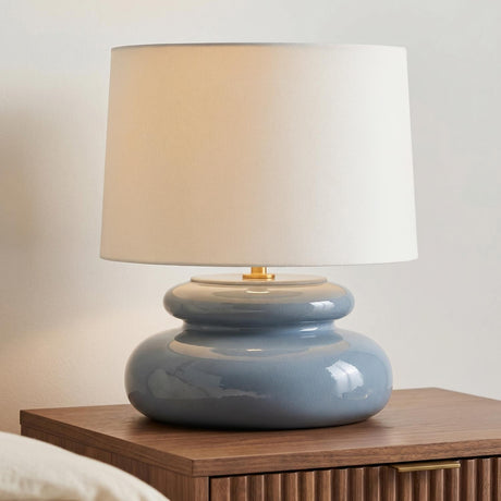 A modern bedroom featuring the Ellsworth Table Lamp 20", Blue Crackle Ceramic with a white linen shade on a fluted walnut nightstand beside a white wall.