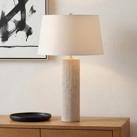 A minimalist living room with a Cassian Table Lamp 30 inch featuring a fluted travertine base and linen shade on a wood console beside black abstract art.