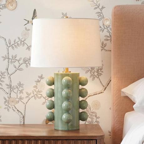 A contemporary bedroom featuring the Tavia Table Lamp in Sage Crackle Ceramic and Aged Brass on a fluted wood nightstand beside floral wallpaper and a pink headboard.