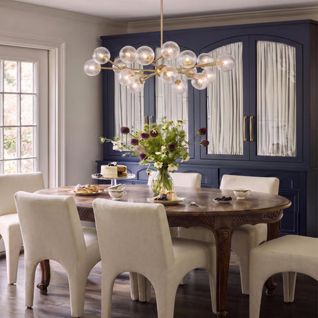 A transitional dining room featuring the Zinnia Chandelier 32-inch in satin brass and clear hammered glass over an ornate wood table and navy blue hutch.