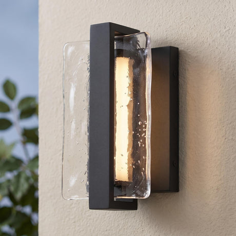 A modern home exterior with textured black Kendrick LED Outdoor Wall Light with seeded cast glass glowing on a beige stucco wall at dusk beside green foliage.