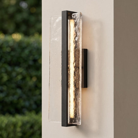 A modern patio setting with a Kendrick LED Outdoor Wall Light in textured black and seeded cast glass glowing on a beige stucco pillar beside green garden hedges.