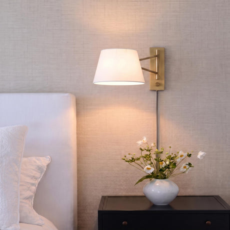 A transitional bedroom with a Barton Medium Swing Arm Wall Sconce in aged brass and white linen mounted beside a white headboard and black nightstand.