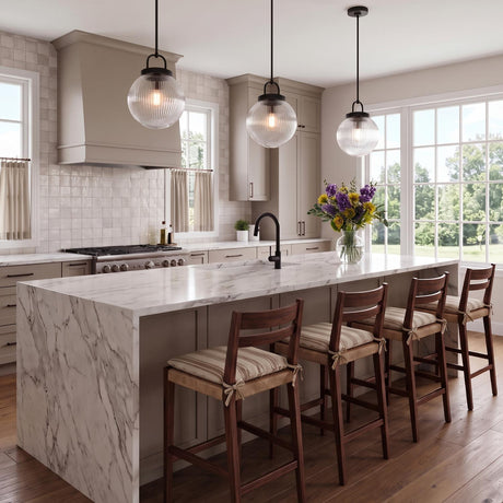 A transitional kitchen with three matte black Whitaker Pendants with clear ribbed glass suspended over a white marble waterfall island featuring greige cabinets and wood barstools.